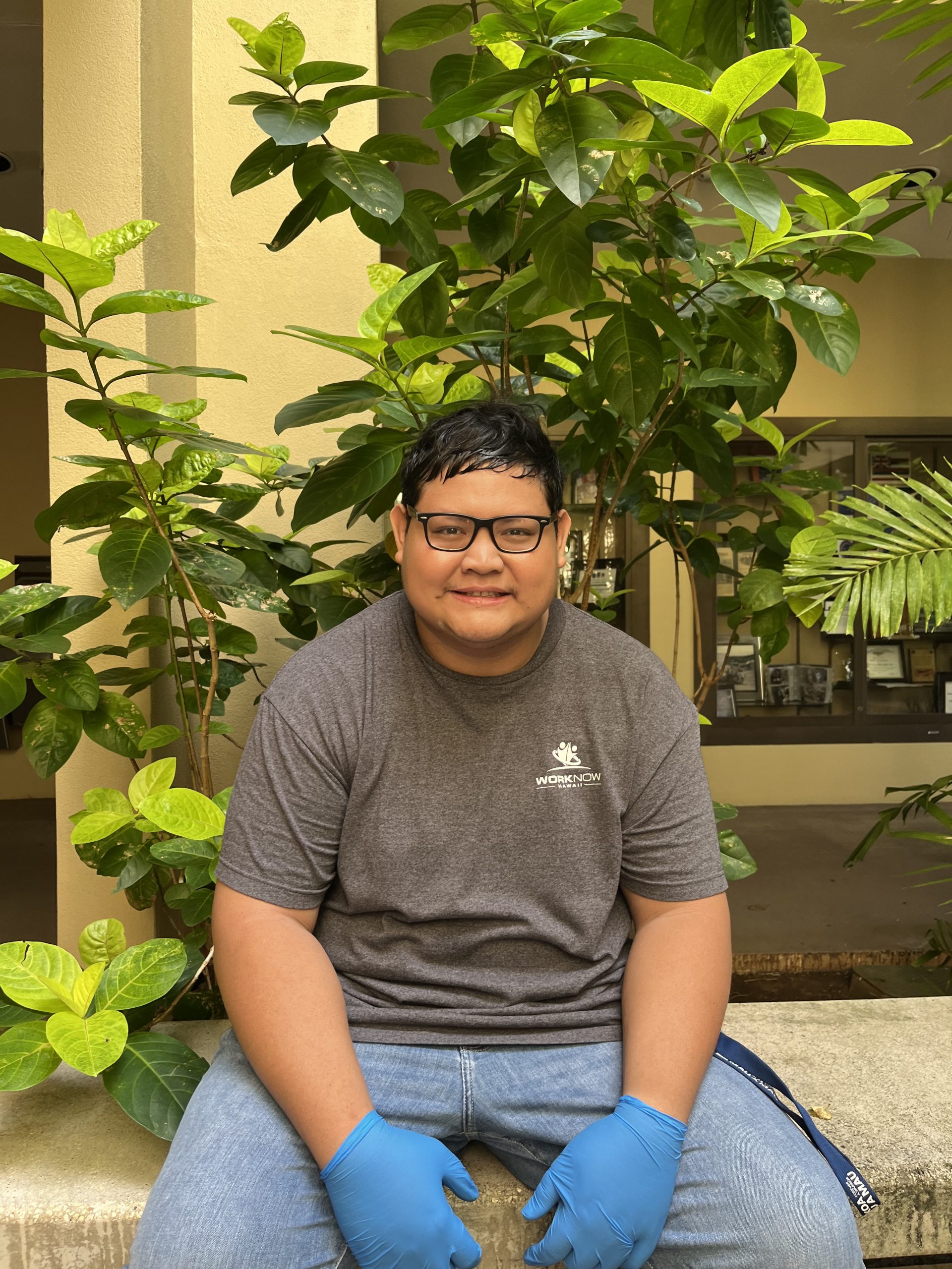 Image of Luke, in his Work Now Hawaii shirt, sitting on a bench in front of a leafy plant.