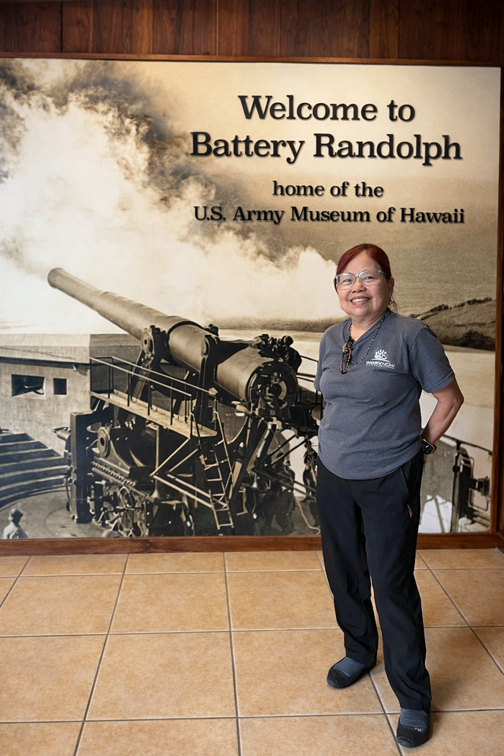 Image of Imelda standing in her Work Now Hawaii T-shirt in front of a giant picture of Battery Randolph. The giant picture has text on top of the top right corner that reads "Welcome to Battery Randolph home of the U.S. Army Museum of Hawaii"