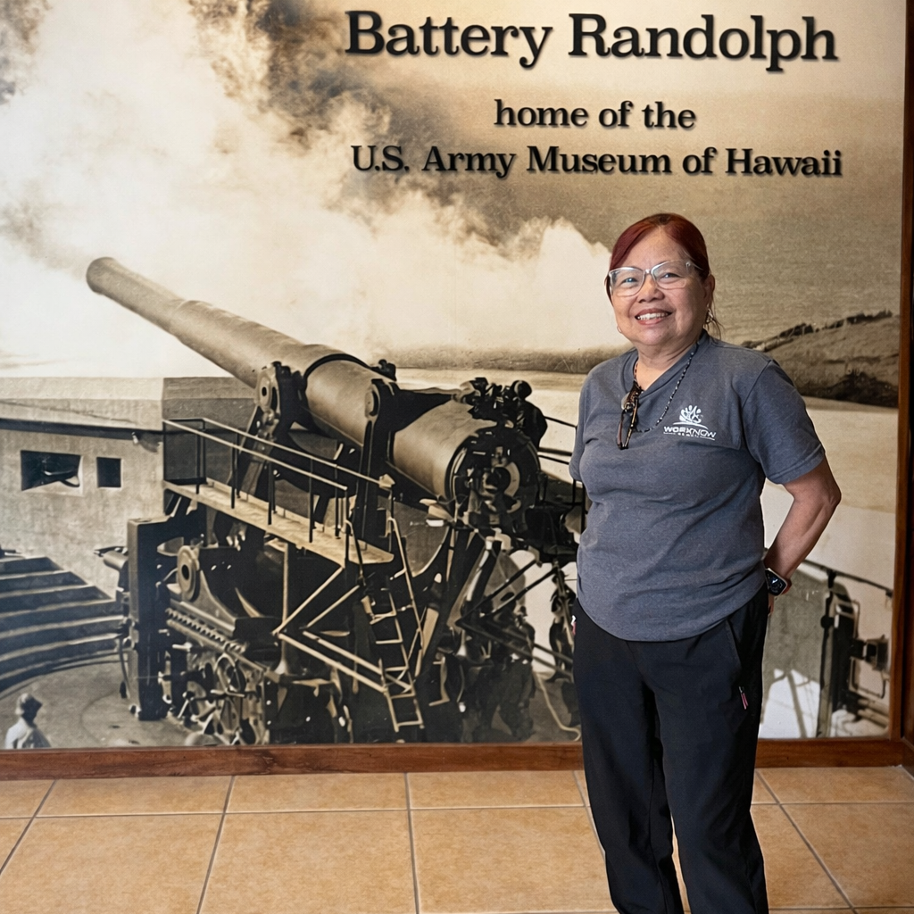 Image of Imelda standing in her Work Now Hawaii T-shirt in front of a giant picture of Battery Randolph. The giant picture has text on top of the top right corner that reads "Welcome to Battery Randolph home of the U.S. Army Museum of Hawaii"
