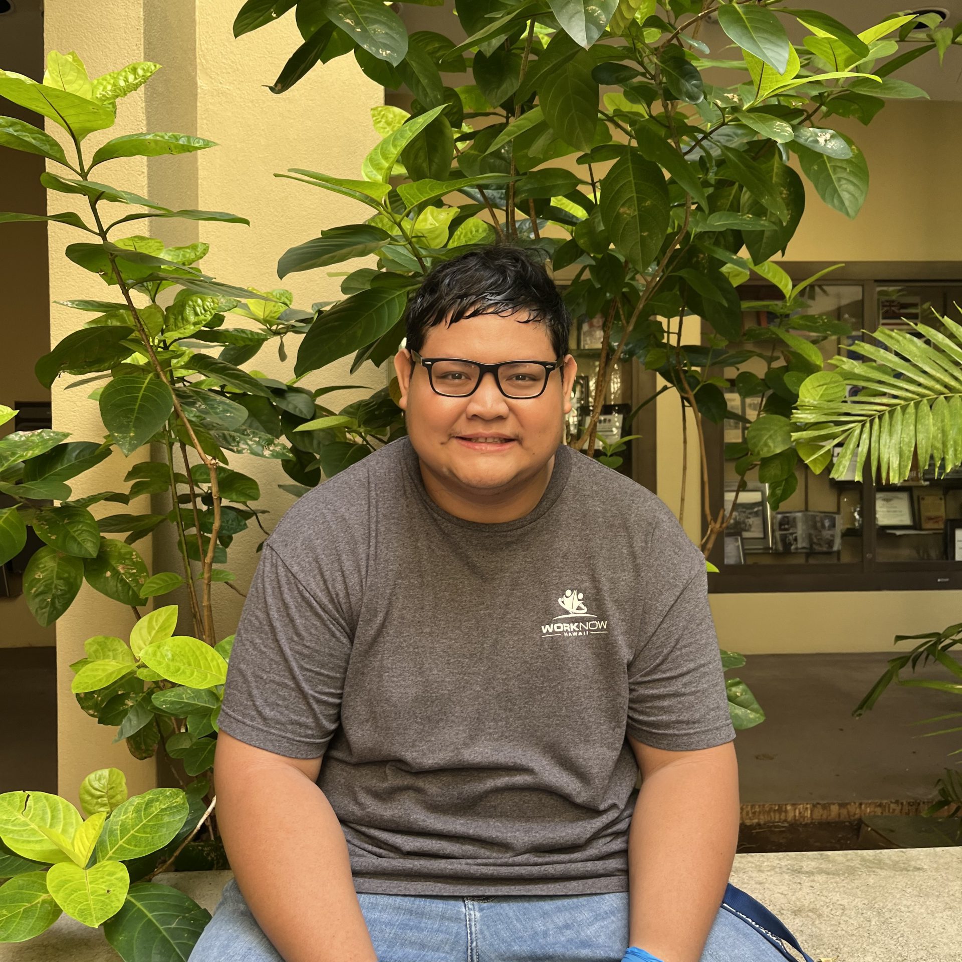Image of Luke, in his Work Now Hawaii shirt wearing sitting on a bench in front of a leafy plant.